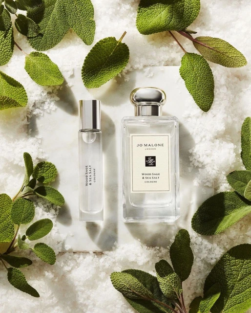 Two Jo Malone Wood Sage & Sea Salt cologne bottles on a marble surface, surrounded by fresh sage leaves and scattered salt granules.