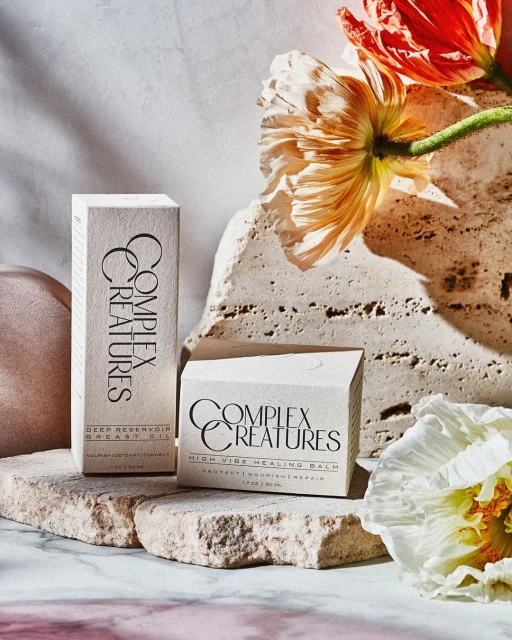 Two 'Complex Creatures' branded skincare product boxes with floral elements arranged on a stone surface, bathed in soft, diffused light.