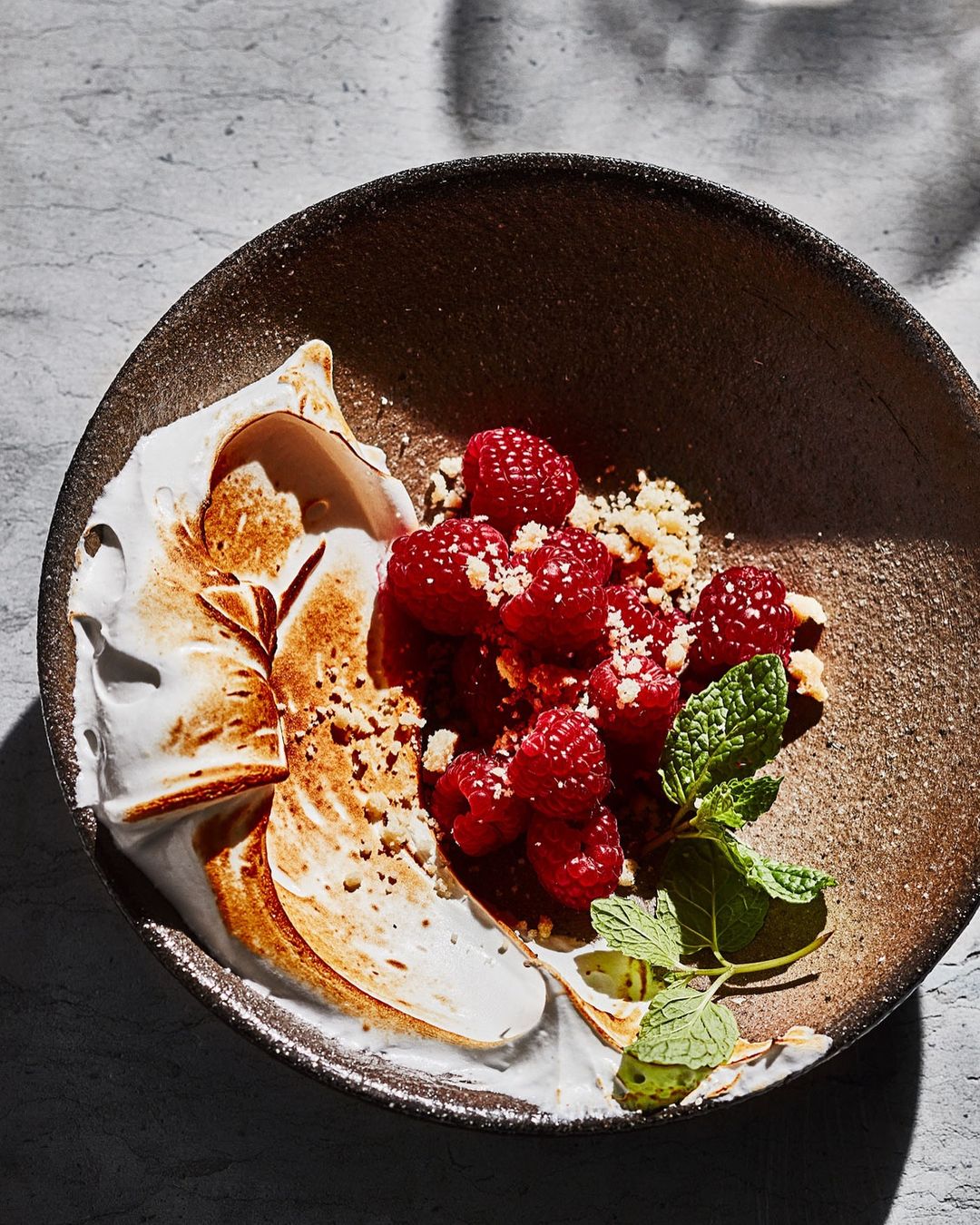 Overhead view of a plate with a dessert featuring raspberries, crumbled toppings, white meringue, and fresh mint garnish.