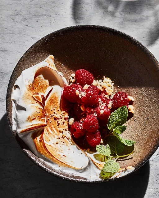 Overhead view of a plate with a dessert featuring raspberries, crumbled toppings, white meringue, and fresh mint garnish.