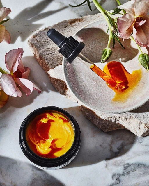 Overhead shot of skincare product jar with dropper on stone surface surrounded by peach colored flowers in a beauty product shot.