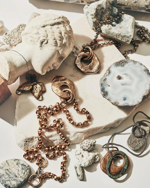 Still life of necklaces and stone objects, including a sculpture bust on a marble surface. Warm, diffused light.