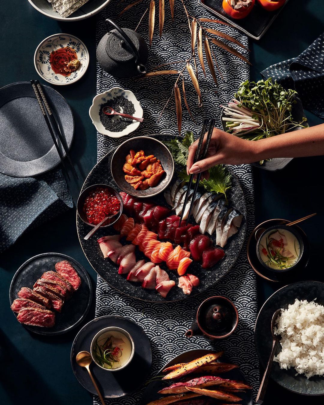Overhead shot of a table laid with various Japanese food items, including sushi, sashimi, rice, and roasted sweet potatoes.