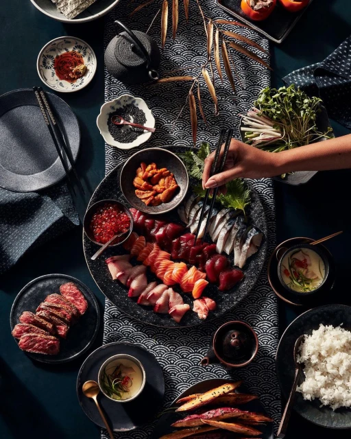 Overhead shot of a table laid with various Japanese food items, including sushi, sashimi, rice, and roasted sweet potatoes.