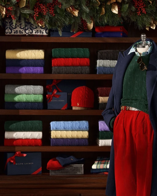 Display of folded cable-knit sweaters and a red knit hat on a wooden shelf, with a mannequin wearing a coat and sweater.
