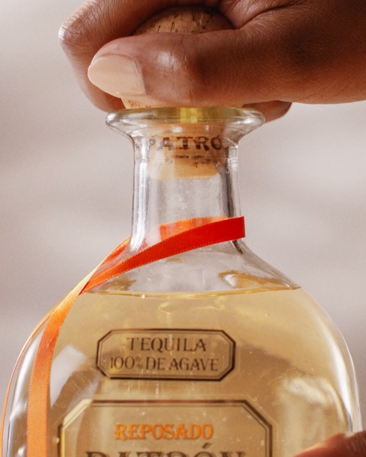 Close-up of a bottle of Patron tequila with someone's hand removing the cork, an orange ribbon, and a neutral background.