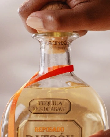 Close-up of a bottle of Patron tequila with someone's hand removing the cork, an orange ribbon, and a neutral background.