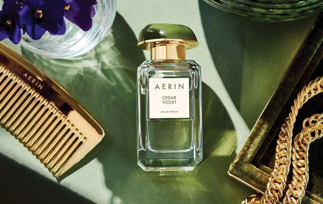 Still life of AERIN Cedar Violet perfume, gold hair comb, and gold chain on green background, accented by purple flowers in glass vase.