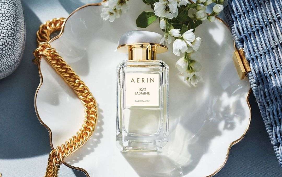 AERIN Ikat Jasmine Eau de Parfum on a decorative white tray with a gold chain, white flowers, and a textured gray vase.