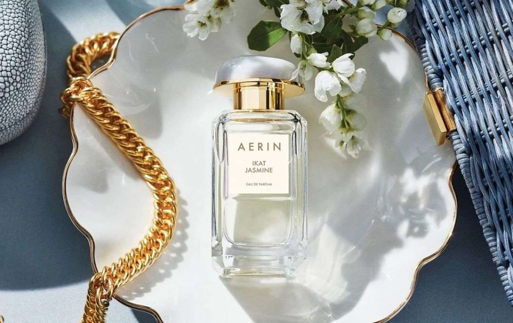 AERIN Ikat Jasmine Eau de Parfum on a decorative white tray with a gold chain, white flowers, and a textured gray vase.