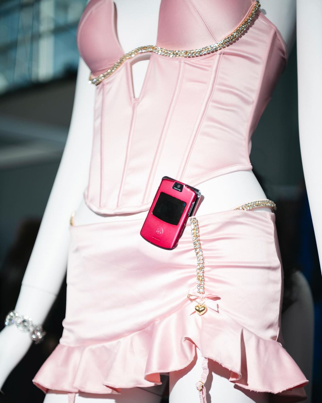 A pink bustier and skirt are displayed on a mannequin, accented with rhinestones and a pink cell phone attached to the waist.