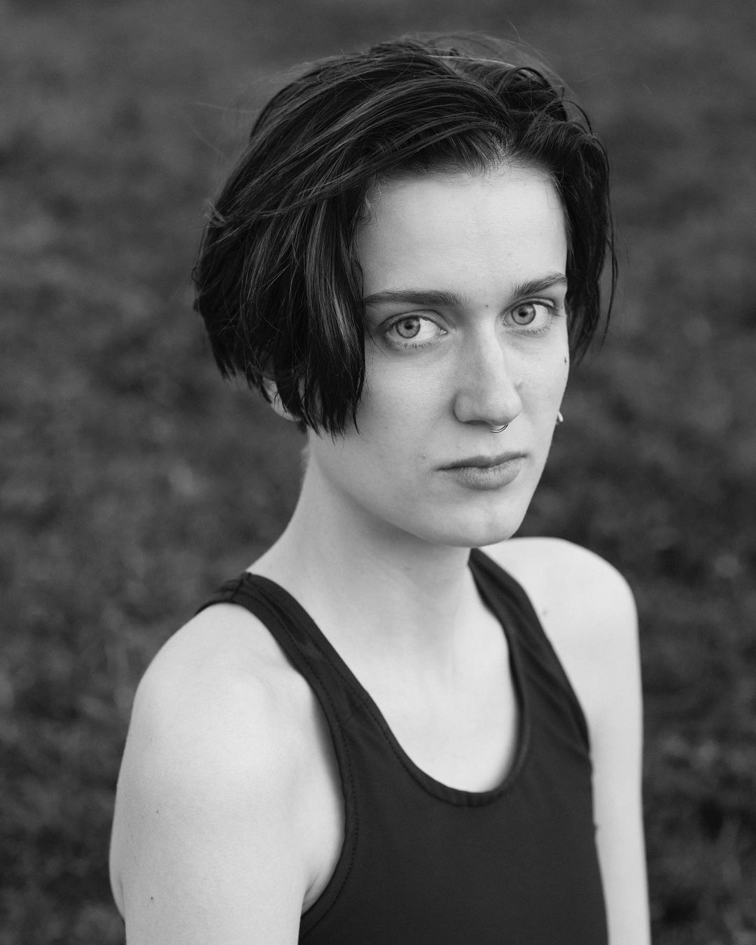 Black-and-white portrait of a young adult female with short dark hair wearing a black tank top, posing outdoors with an intense stare.