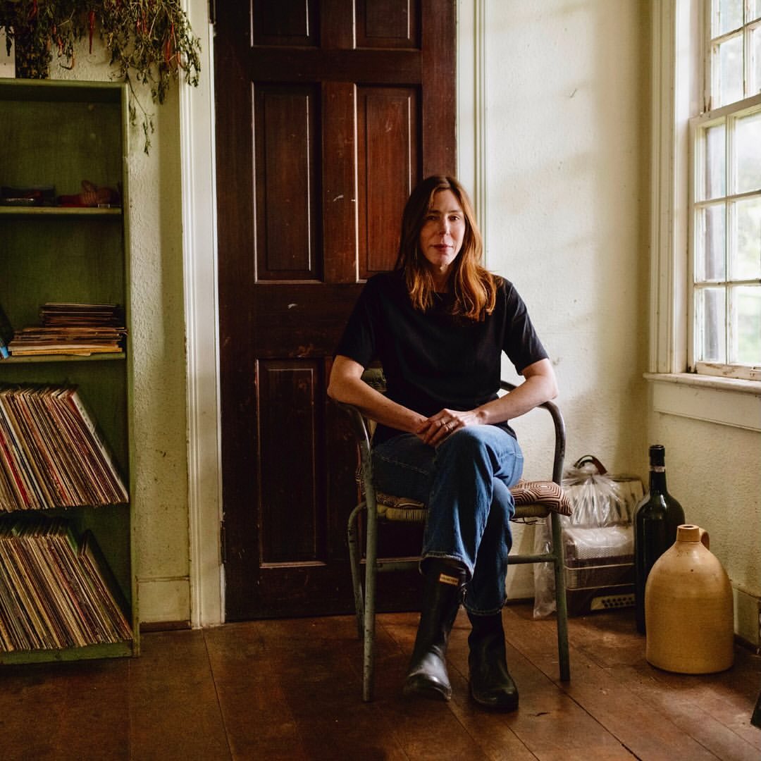 Woman with auburn hair sits in a chair indoors near a window and a wood door. Vintage elements and neutral colors create a cozy scene.