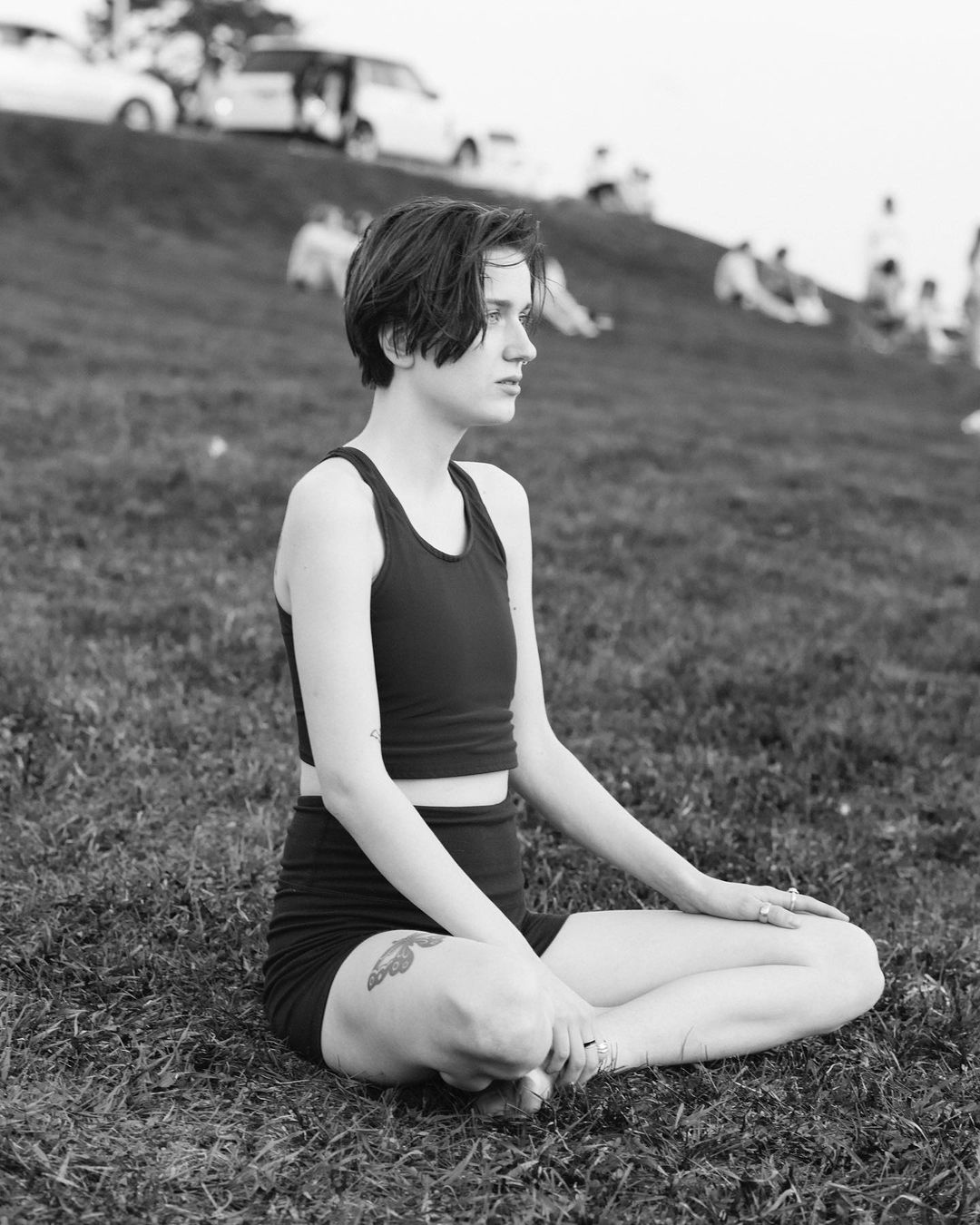 Young woman with short dark hair in black athletic clothing sits cross-legged on grass in a monochromatic outdoor portrait.