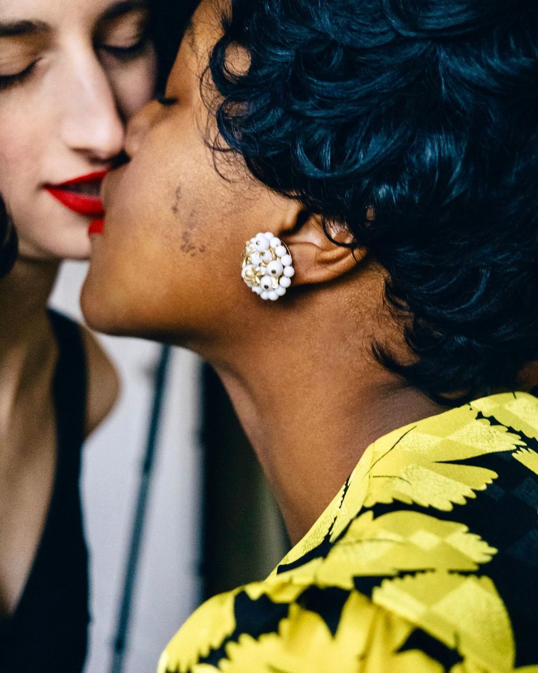 Close-up of two adult females, one Black and one Caucasian, face to face; the Black female has an elaborate pearl earring and yellow floral top.