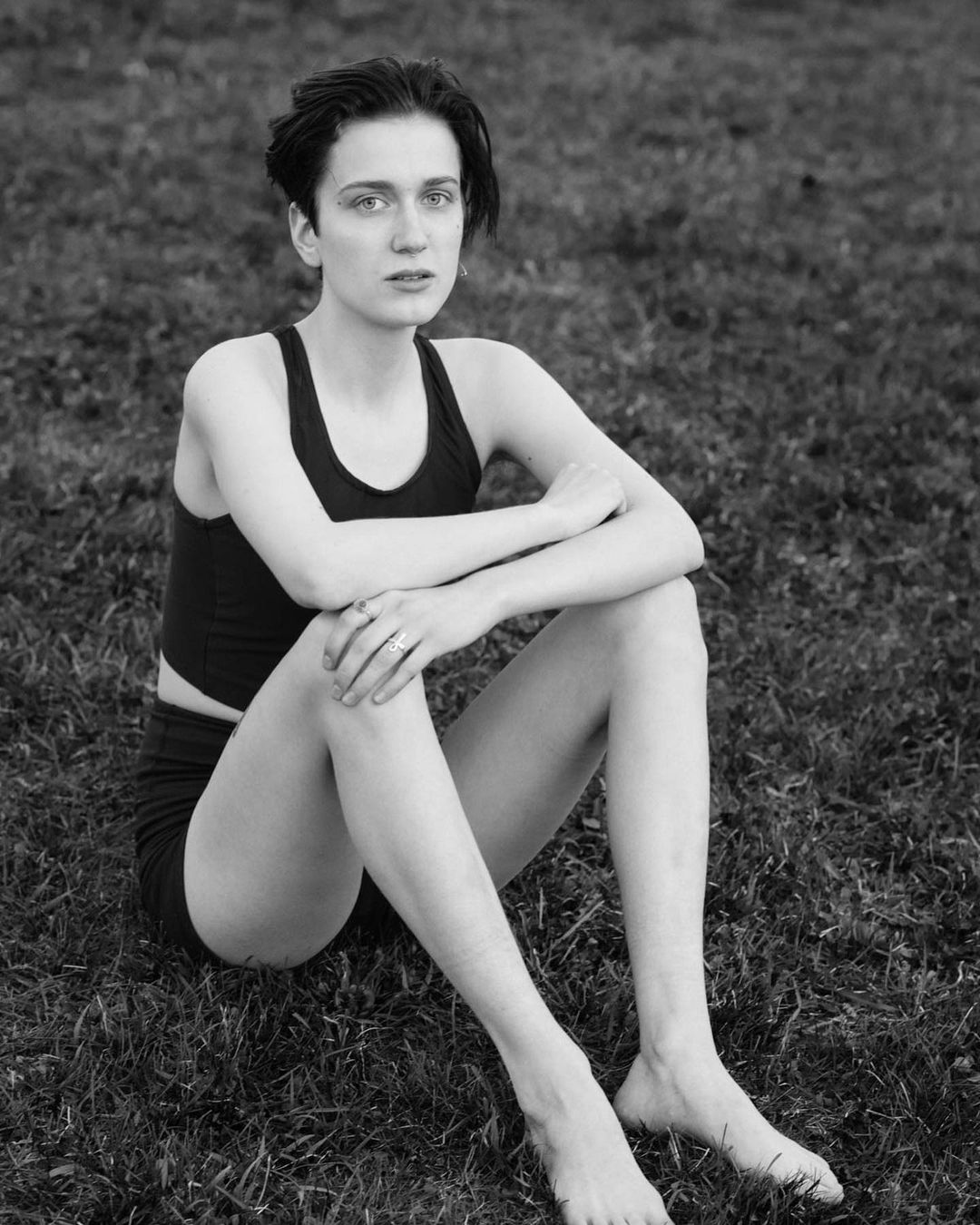 Black and white portrait of a young woman sitting cross-legged in a grassy field, wearing a tank top and shorts, with a serious expression.