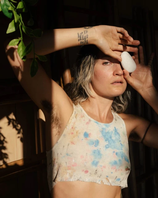 An adult woman holds a stone to her face in an indoor setting, lit by natural light. The woman has wavy hair and is wearing a tie-dye top.