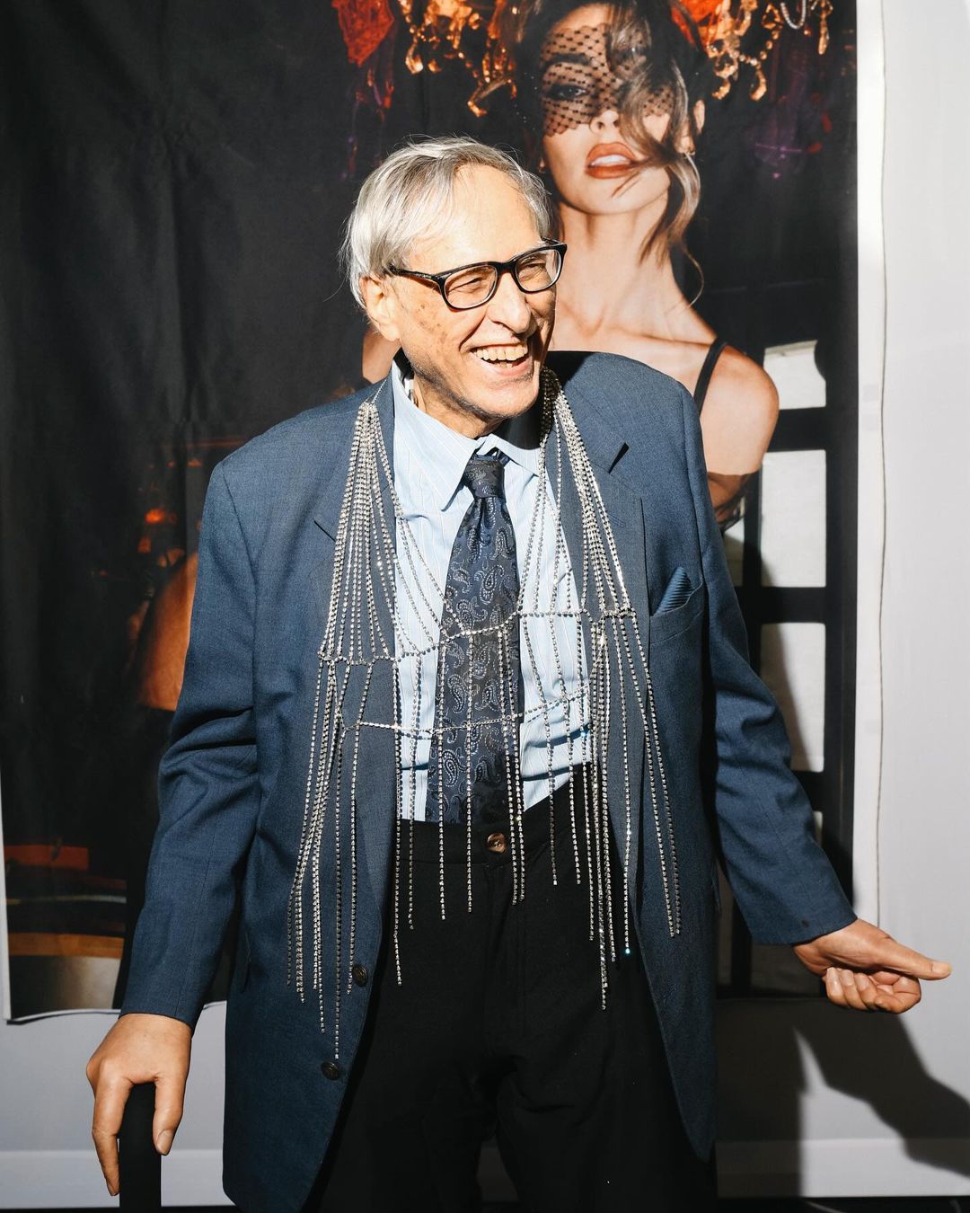 Elderly man in a blue suit and glasses smiles next to a poster of a model with a face covering and dark clothing. 