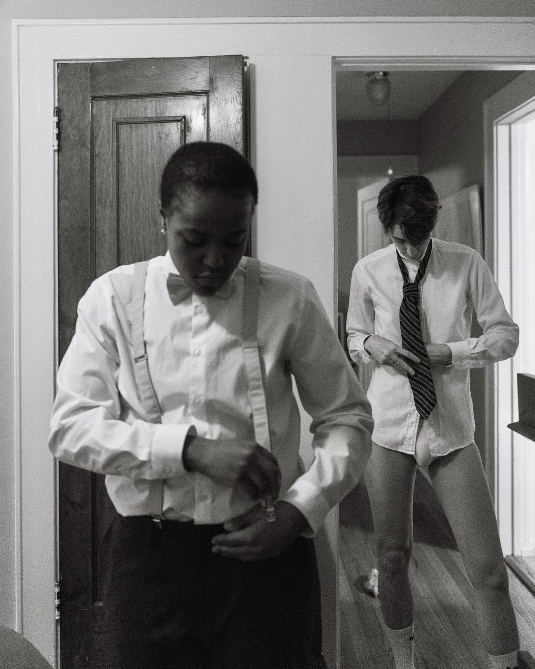 Monochrome portrait of two people in a room: one in a bow tie and suspenders, the other adjusting a tie, with doors in the background.