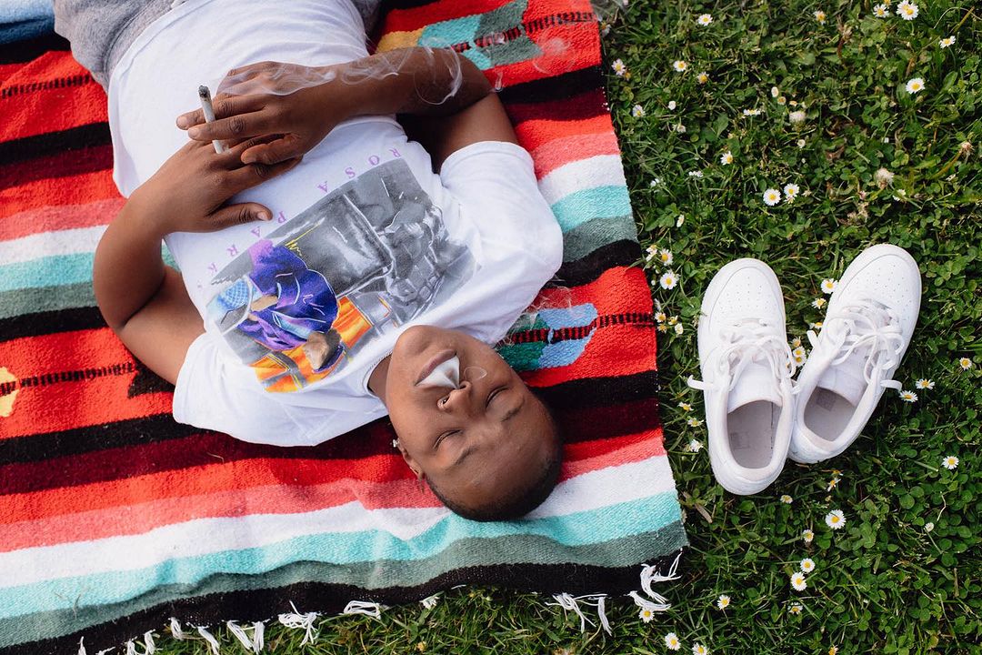 Woman lounges on a striped blanket outdoors on green grass with white sneakers, holding a cigarette with smoke rising in the air.