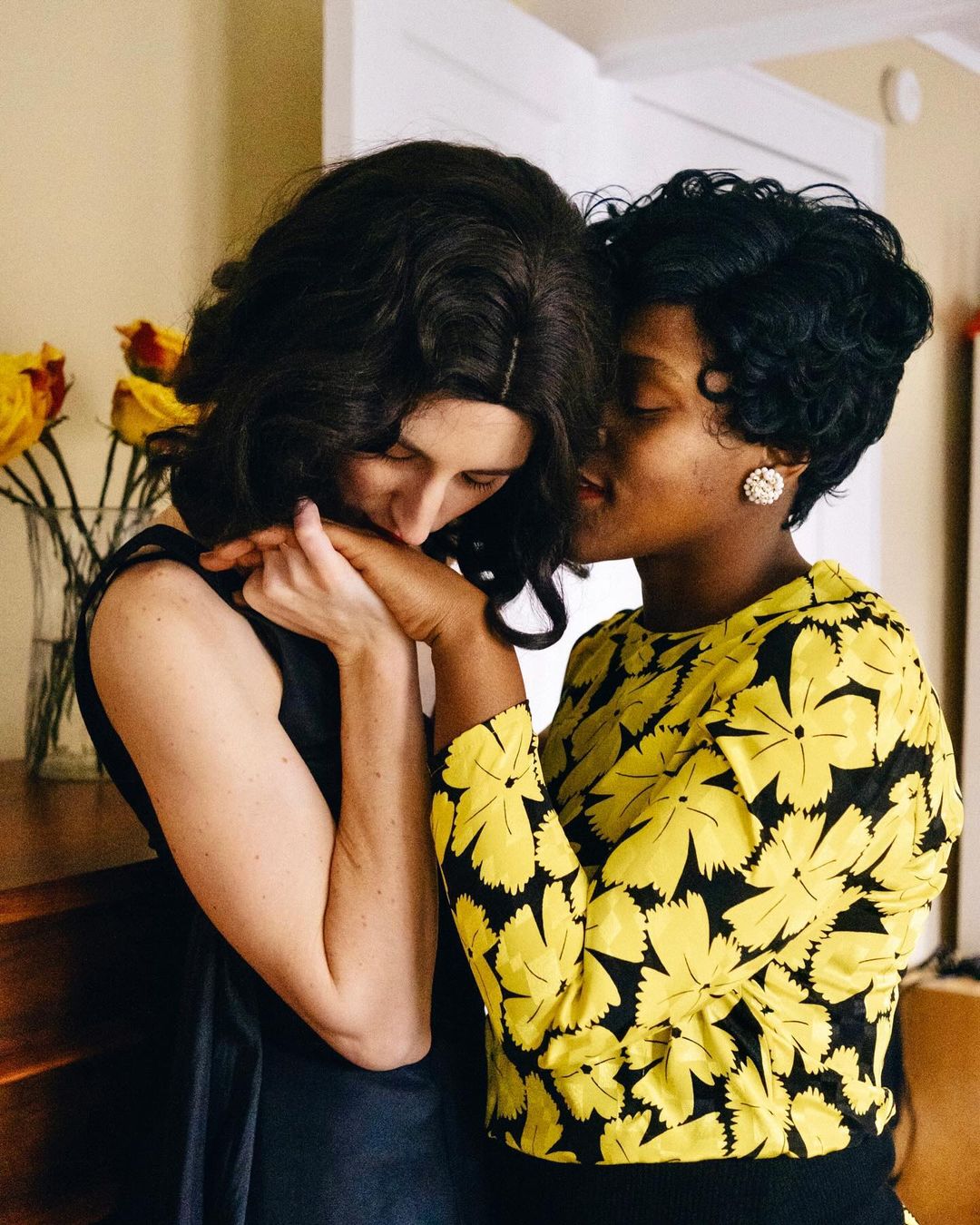 Two women in vintage-style dresses embrace indoors; one rests her head near the other's hand, roses in a vase are visible.