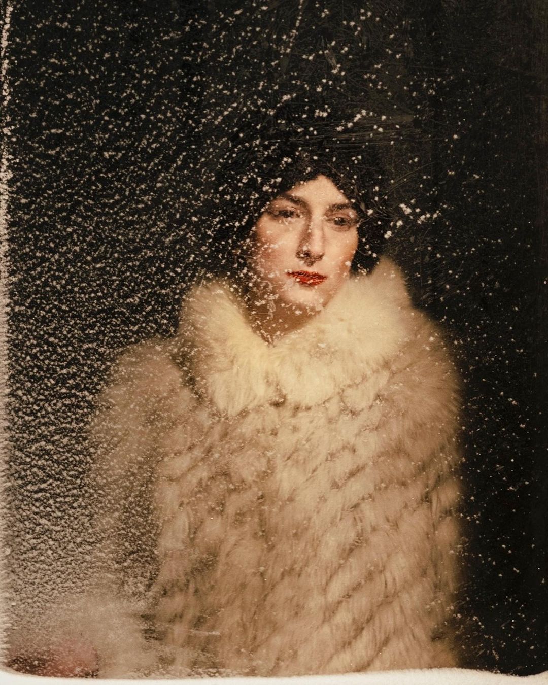 Elegant woman in fur coat stands behind frosted glass, exhibiting a blend of fashion and artistic photography in a dark setting.