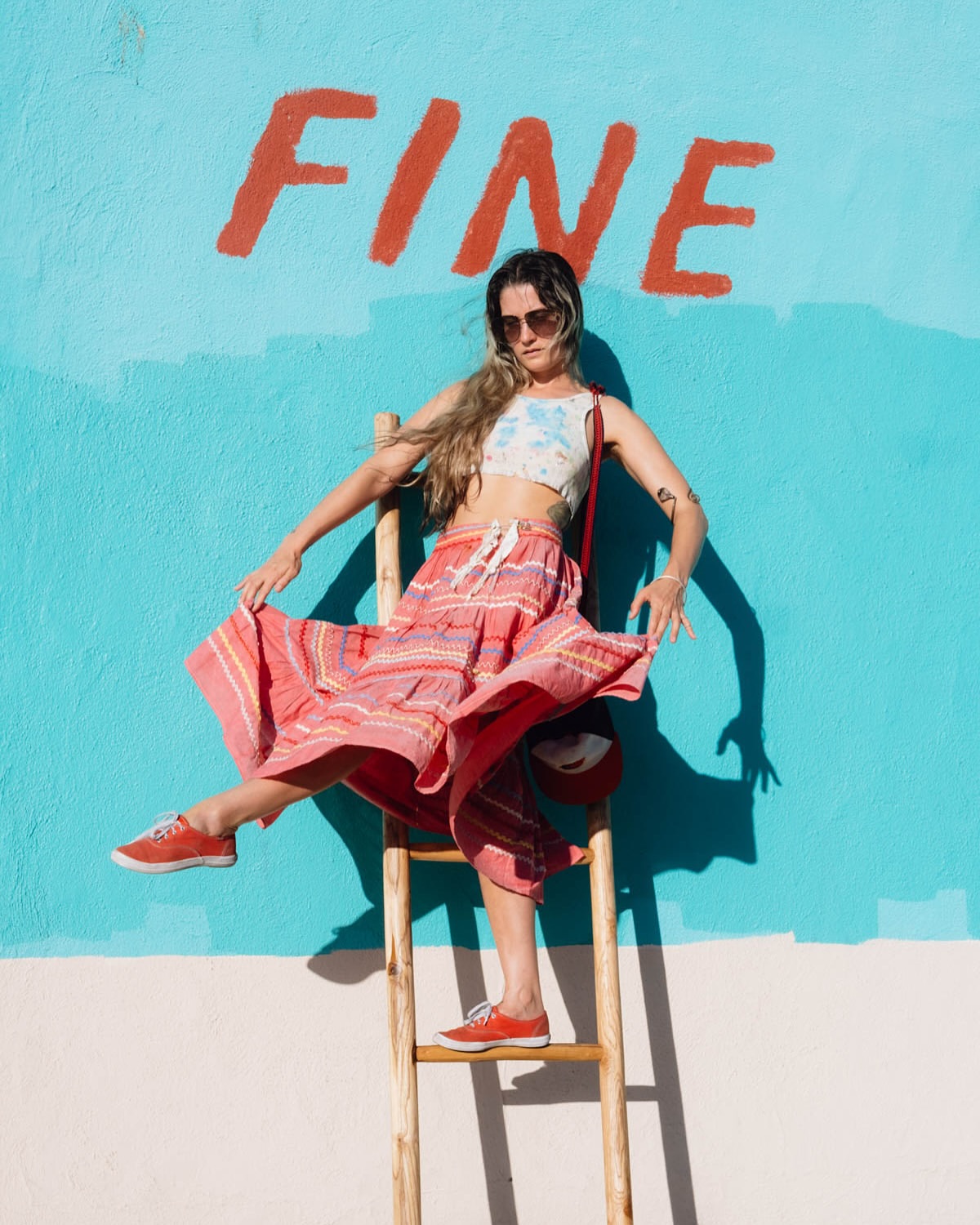 A woman in a flowing skirt sits on a ladder against a blue and white wall with the word 'FINE' painted on it in red.