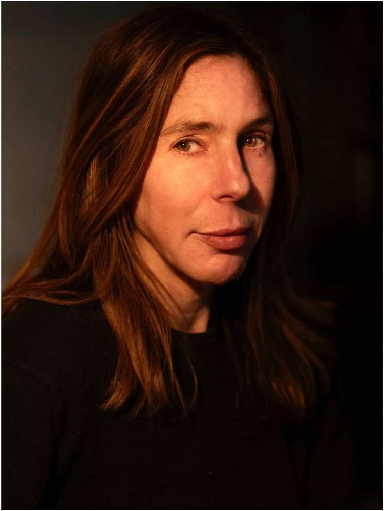 Close-up portrait of an adult woman with long auburn hair wearing a black top, illuminated by directional light in a dark, indoor setting.