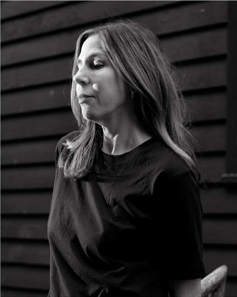 Black and white portrait of a woman with shoulder-length hair, wearing a dark top, against a dark, horizontal-lined background.