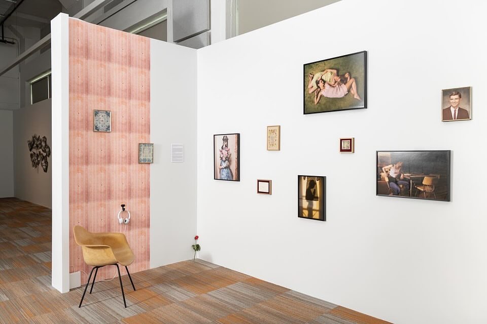 Art gallery exhibit featuring framed pictures on a white wall, a wooden chair, pink wallpaper, and a polished floor. Interior design.