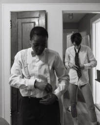 Black-and-white portrait of two individuals dressing indoors, one in foreground adjusting suspenders.