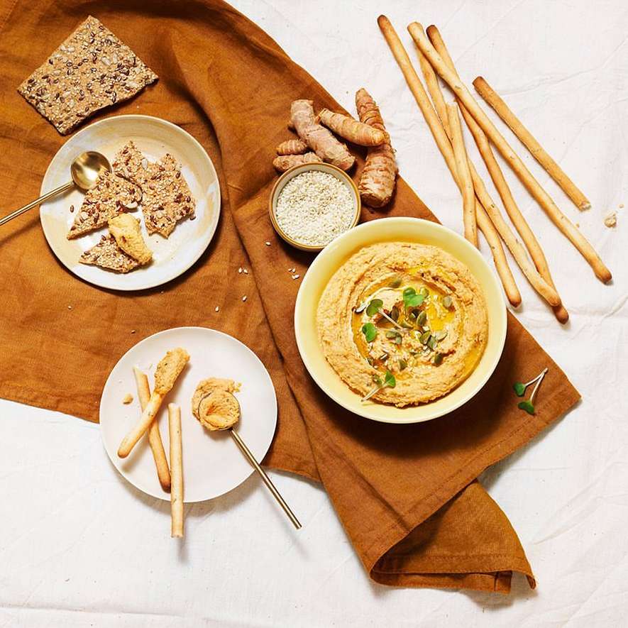Overhead shot of hummus served in a yellow bowl alongside turmeric, flaxseed crackers, and breadsticks on a rustic brown tablecloth.