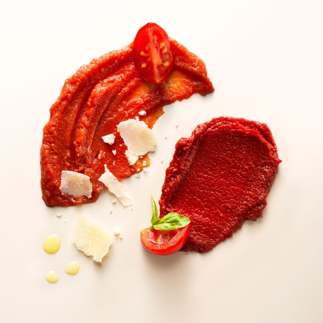 Overhead food photo of tomato sauce smears with halved tomatoes, basil, and cheese shavings on a creamy white background, studio lit.