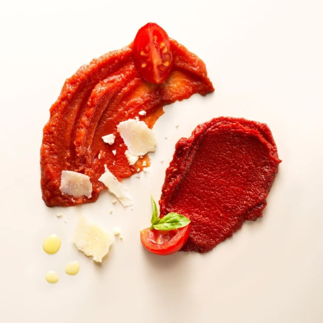Overhead food photo of tomato sauce smears with halved tomatoes, basil, and cheese shavings on a creamy white background, studio lit.