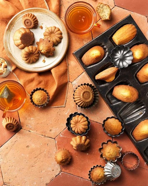 Overhead shot of madeleine cookies in baking pan and on plate, accompanied by cups of tea on orange tile surface.