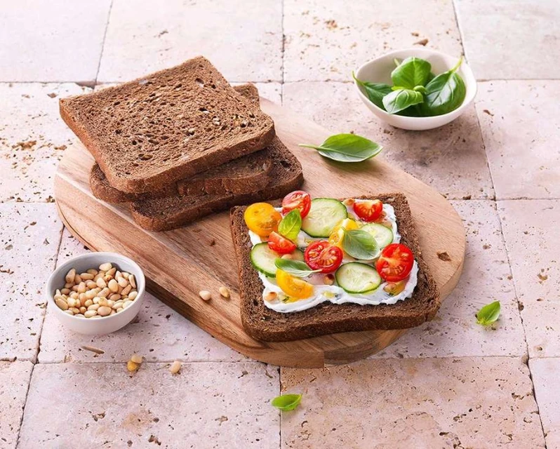 Dark rye bread slices and an open-faced sandwich with cream cheese, cucumbers, tomatoes, and basil on a wooden board.
