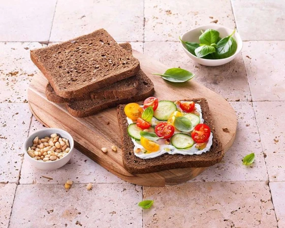 Dark rye bread slices and an open-faced sandwich with cream cheese, cucumbers, tomatoes, and basil on a wooden board.