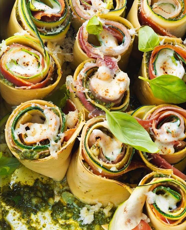 Overhead shot of zucchini roll-ups with ham, cheese, and pesto sauce, arranged on a platter. The dish is garnished with fresh basil leaves.