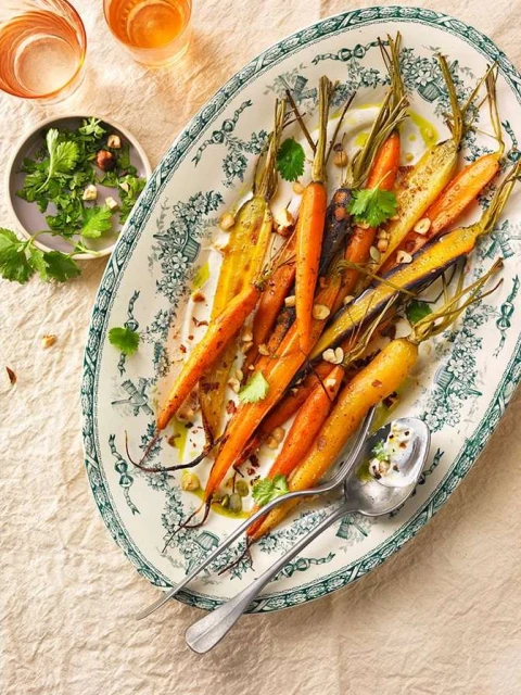 Overhead shot of roasted carrots on an ornate platter, garnished with nuts and herbs, with drinks in glasses on a beige tablecloth.