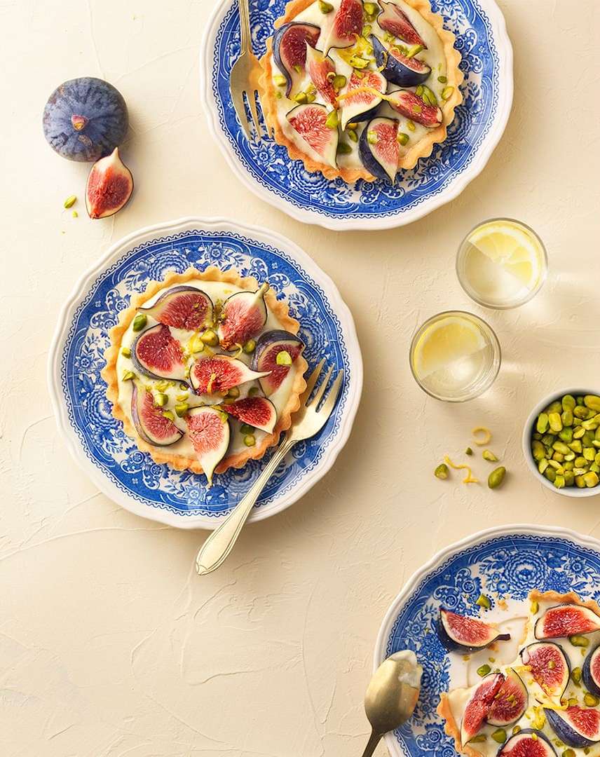 Overhead shot of three fig tarts on patterned blue and white plates, garnished with pistachios, accompanied by drinks and a bowl of pistachios.