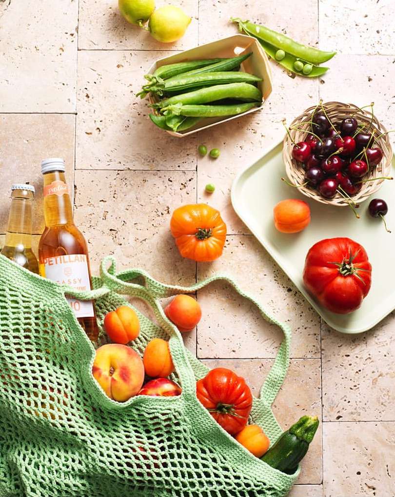 Overhead shot of a green string bag with peaches, apricots, tomato, zucchini, and bottles, surrounded by fruit and vegetables on tiles.