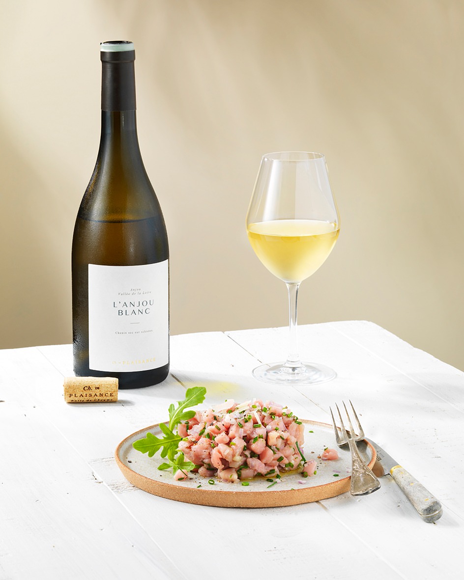 Still life of tuna tartare with greens on a plate next to a wine bottle, wine glass, and cork, all on a white wooden surface.