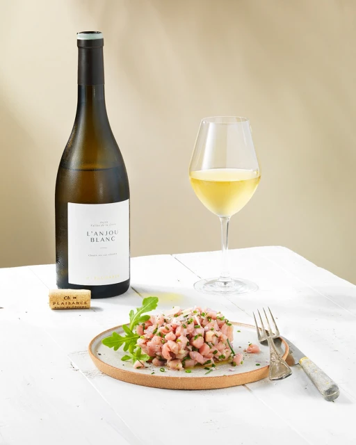 Still life of tuna tartare with greens on a plate next to a wine bottle, wine glass, and cork, all on a white wooden surface.