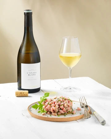 Still life of tuna tartare with greens on a plate next to a wine bottle, wine glass, and cork, all on a white wooden surface.