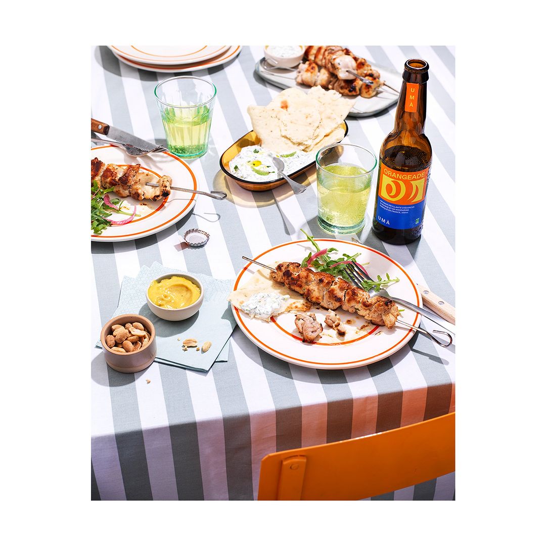 Overhead shot of a table laid out with grilled souvlaki skewers on plates, dips, flatbread, beverages, and peanuts, on a striped tablecloth.
