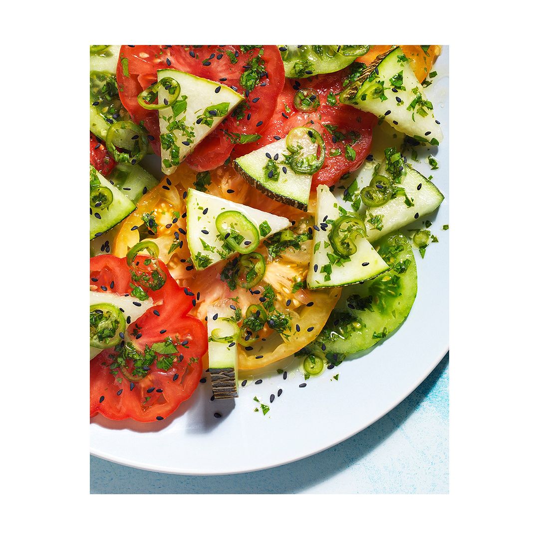 Overhead shot of a colorful heirloom tomato salad with cucumber slices, herbs, and spices, presented on a white plate with bright lighting.