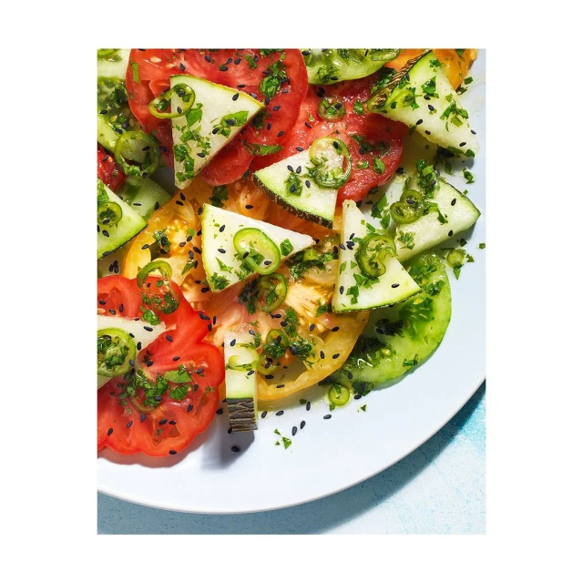 Overhead shot of a colorful heirloom tomato salad with cucumber slices, herbs, and spices, presented on a white plate with bright lighting.