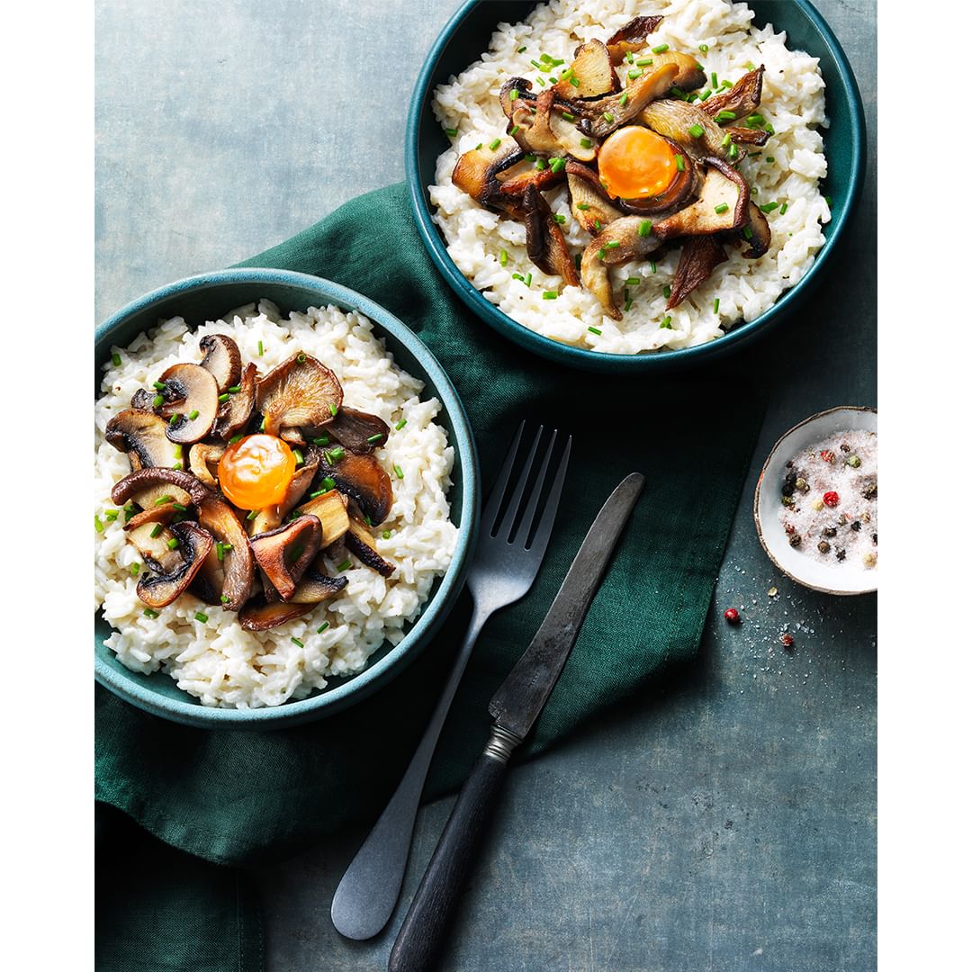 Two bowls of rice topped with fried mushrooms and egg yolk are placed on a table with a knife, fork, and green napkin.