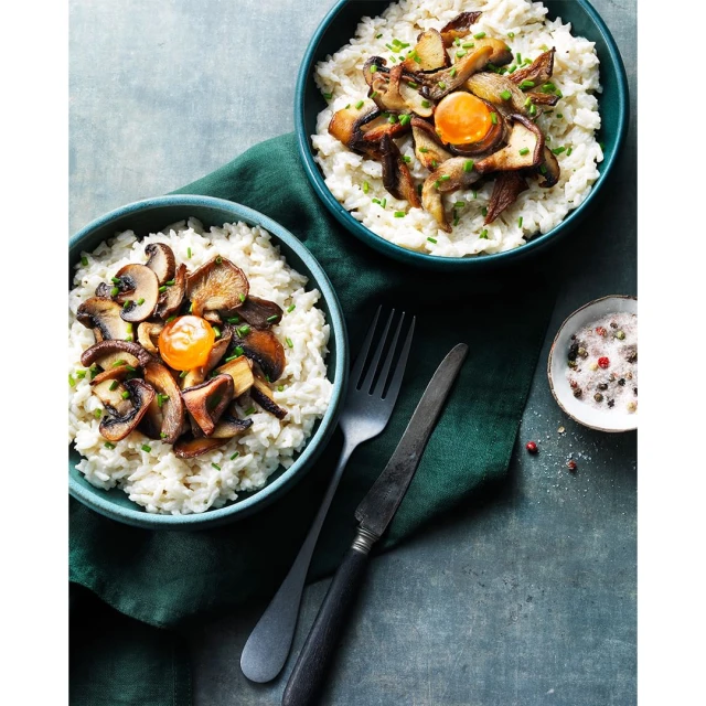 Two bowls of rice topped with fried mushrooms and egg yolk are placed on a table with a knife, fork, and green napkin.