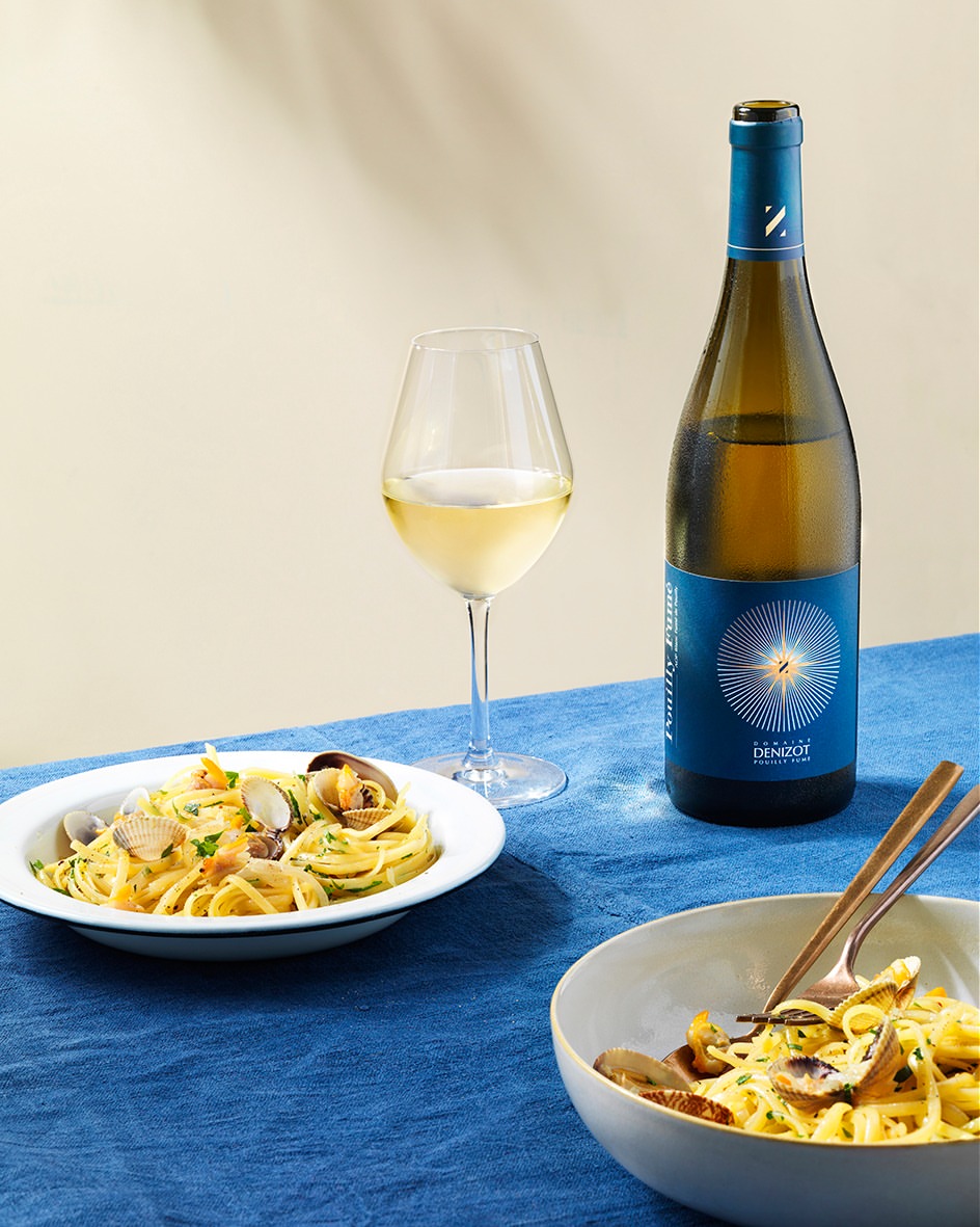 A bottle of white wine is situated near a plate of spaghetti with clams and another pasta bowl on a blue tablecloth.